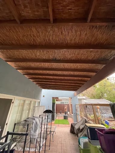 Covered patio with woven reed pergola ceiling, metal chairs, potted plants, and view to backyard